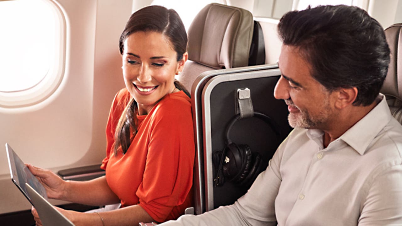 Fotografia de um homem e de uma mulher sorridentes sentados abordo de um avião TAP em classe Executiva. A mulher, morena, usa o cabelo apanhado, encontra-se vestida com uma blusa vermelha  e segura um tablet para o qual está a olhar.  O Homem, moreno, com barba branca, veste uma camisa branca e está a olhar para o tablet que a mulher segura.