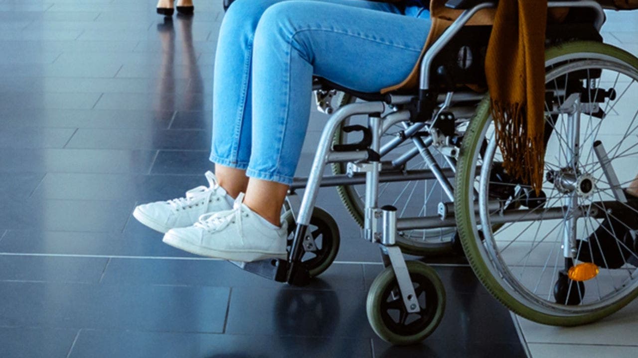 Fotografia de uma mulher a ser transportada numa cadeira de rodas no aeroporto com a ajuda de outra mulher. Do lado esquerdo, uma mala de viagem amarela é puxada por um homem.