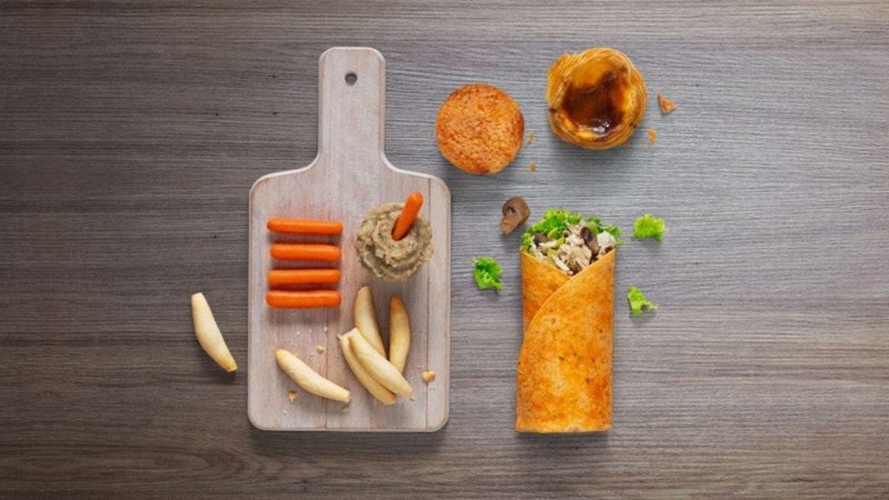 Fotografia de uma mesa de madeira com 4 elementos: uma tábua com cenoura, palitos de pão e uma taça com molho, um wrap de frango, cogumelos e alface, um pastel de feijão e um pastel de nata.