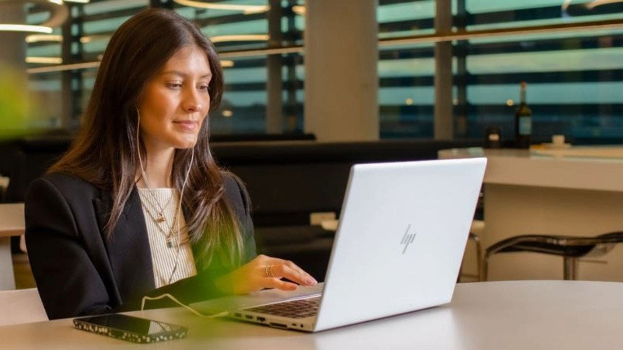 Fotografia de uma mulher caucasiana, com cabelo preto comprido, sentada numa mesa de um Lounge da TAP. A senhora tem uns auriculares nos ouvidos e está com a sua mão direita a mexer num computador portátil cinzento que se encontra sobre a mesa à sua frente. Ainda sobre a mesa, do lado esquerdo do portátil, está um telemóvel. Em primeiro plano da imagem , à esquerda, é possível ver as folhas verdes de uma planta e ao fundo da imagem encontra-se uma janela com vista sobre o aeroporto.