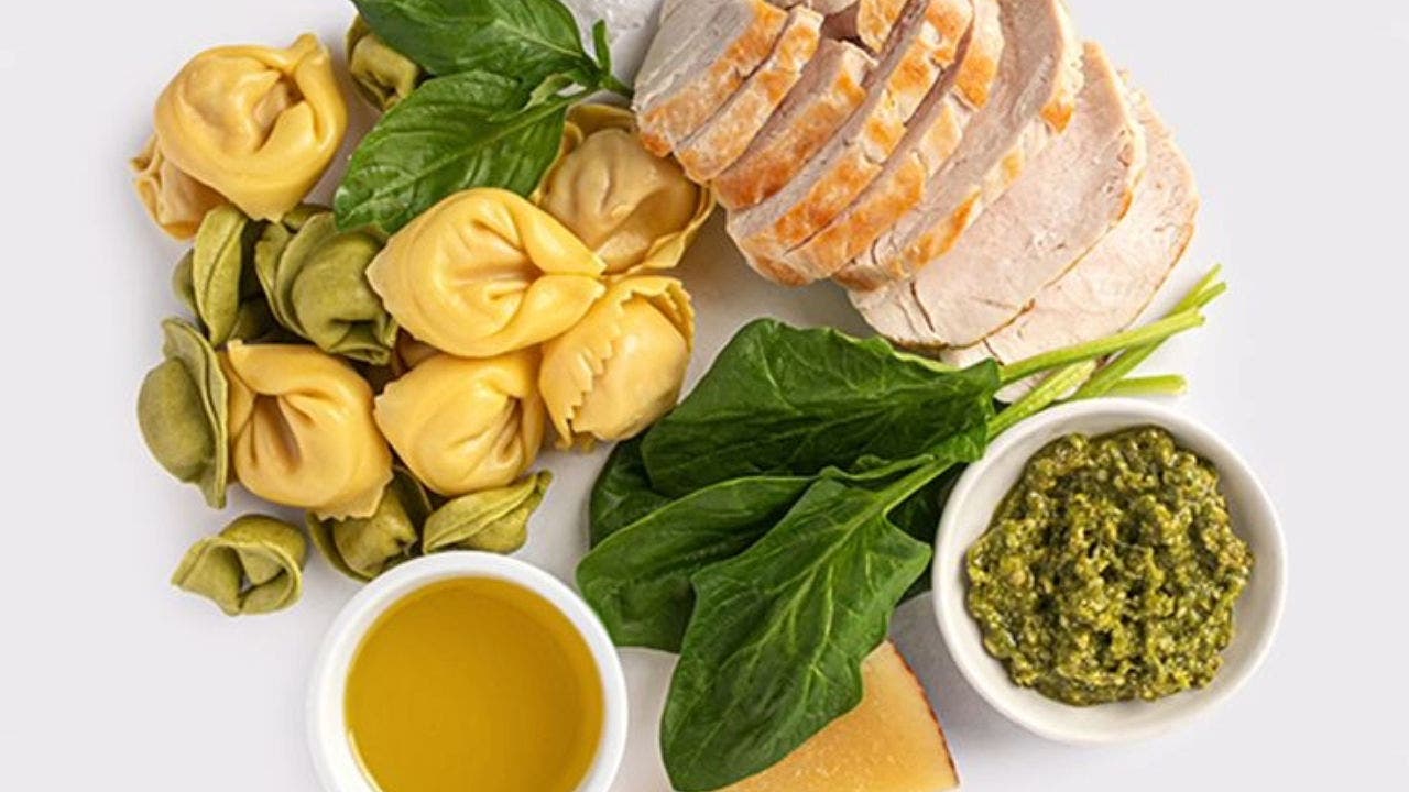 Composição com vários alimentos: em baixo, uma pequena taça com azeite,  uma fatia de queijo, alguns espinafres cruz e uma taça com pesto. Por cima, massas recheadas (de cor amarela e verde) duas folhas de manjericão e carne lombo fatiada.