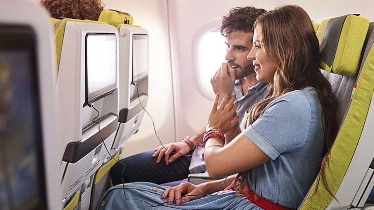 Um casal de passageiros, sentado nas cadeiras verdes da Classe Económica, num ambiente com muita luz natural. Estão curiosos ao assistir um programada de TV a bordo, no ecrã da cadeira da passageira, com os headphones de ouvido colocados.