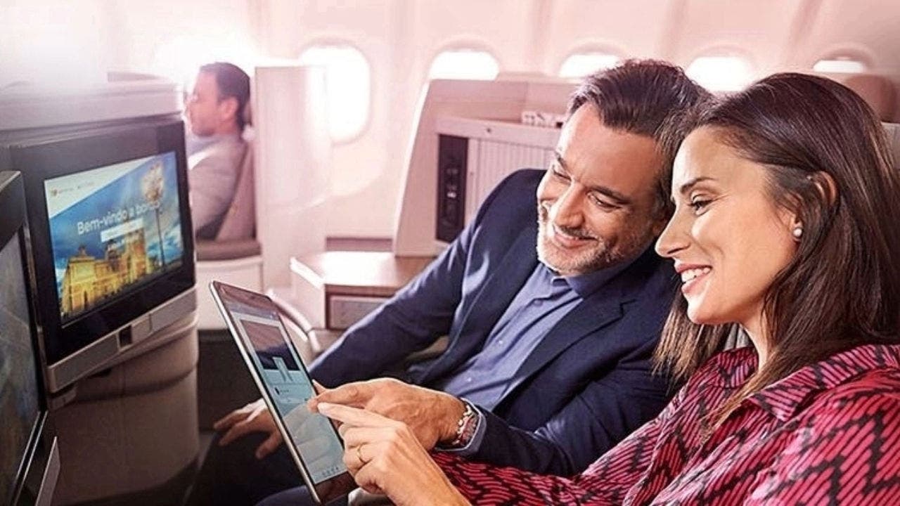 Plano Aproximado de uma mulher e de um homem, sorridentes, sentados a bordo de um avião. A mulher está a segurar um tablet e o homem a olhar para o mesmo. Ambos apontam para o ecrã do tablet. Á sua frente estão dois ecrãs incorporados nos ecrãs nos bancos do avião.