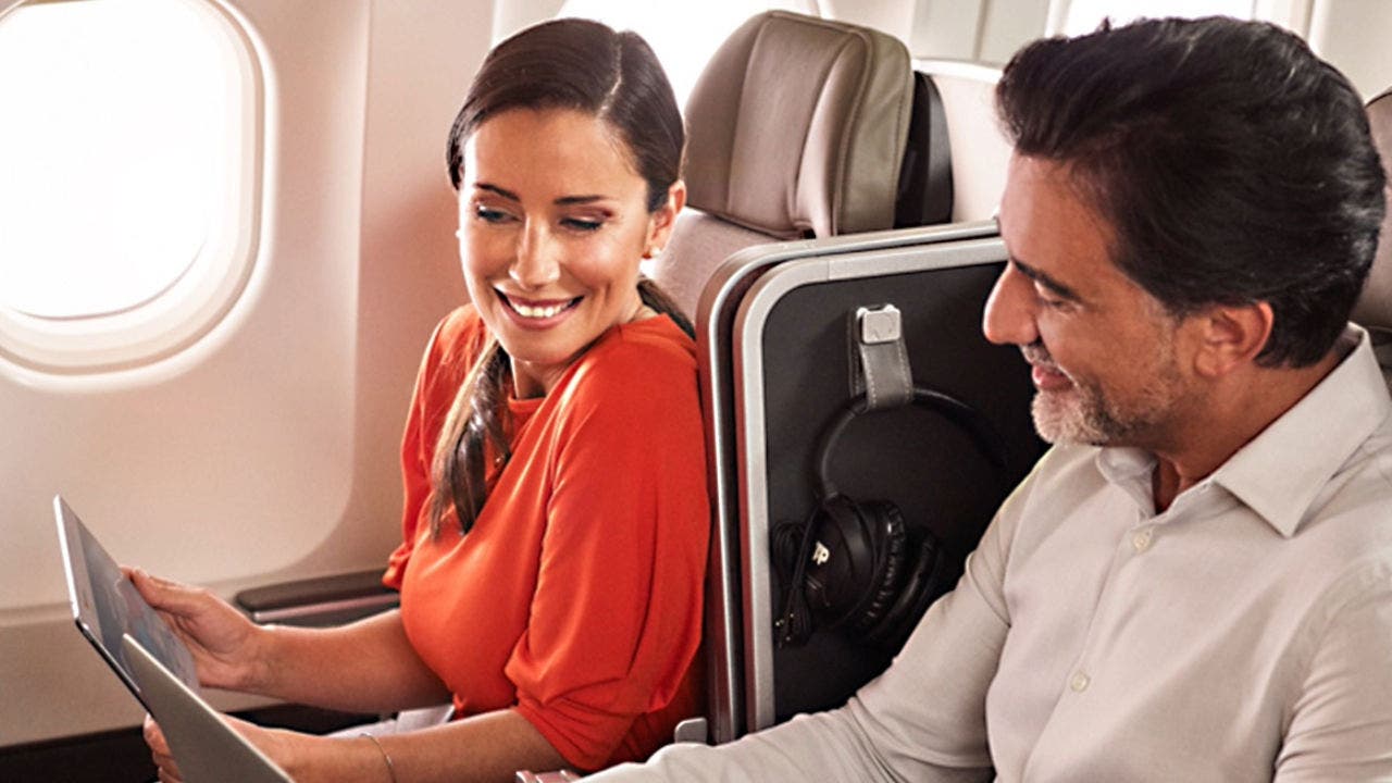 Fotografia de um homem e de uma mulher sorridentes sentados abordo de um avião TAP em classe Executiva. A mulher, morena, usa o cabelo apanhado, encontra-se vestida com uma blusa vermelha e segura um tablet para o qual está a olhar.  O Homem, moreno, com barba branca, veste uma camisa branca e está a olhar para o tablet que a mulher segura.