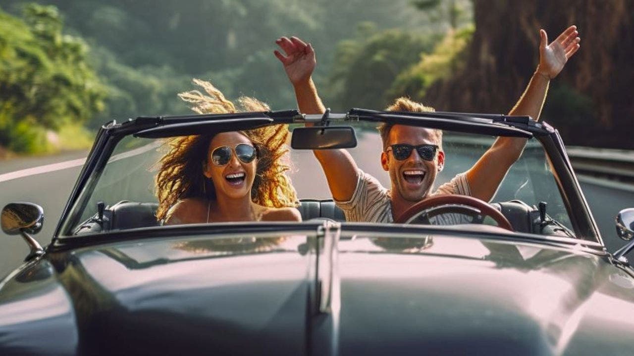 A imagem mostra um homem e uma mulher, sorridentes dentro de um carro cinzento descapotável. O homem vai no lugar do condutor e vai com os braços no ar, a mulher está ao seu lado, no lugar do passageiro. Ambos estão com óculos de sol. Á sua volta rodeia-se uma paisagem de natureza que sugere uma zona de montanhas.